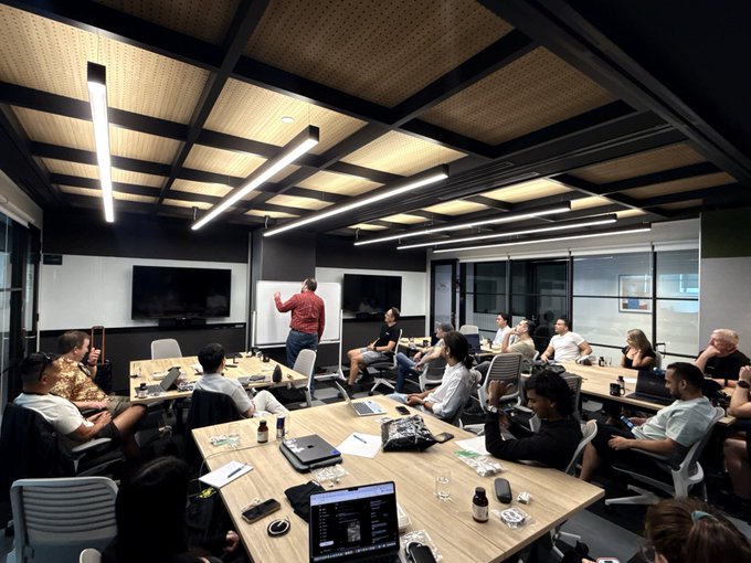 A group of individuals sits in a conference room with wooden ceiling beams and long light fixtures, listening to a man in a red shirt standing at the front, with laptops, monitors, and drinks scattered across the tables.