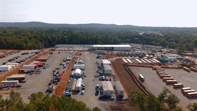 An aerial view of a large outdoor event site or industrial yard with multiple vehicles, trailers, and equipment staged across a cleared area near a tree line.