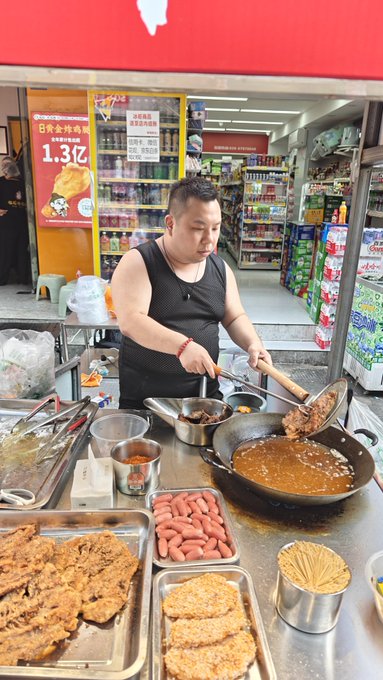 A street food vendor in a black tank top cooks at a stall in front of a convenience store, frying battered meat patties and skewers in a wok with trays of sausages and skewers displayed on the counter.