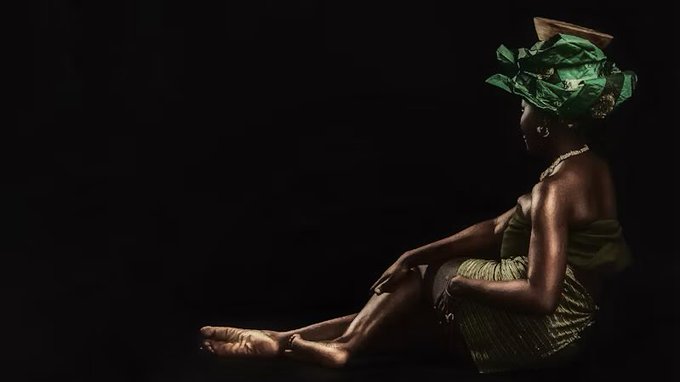 An artistic photograph of a dark-skinned woman seated against a black background, wearing a green wrap and an elaborate green headwrap adorned with jewels.