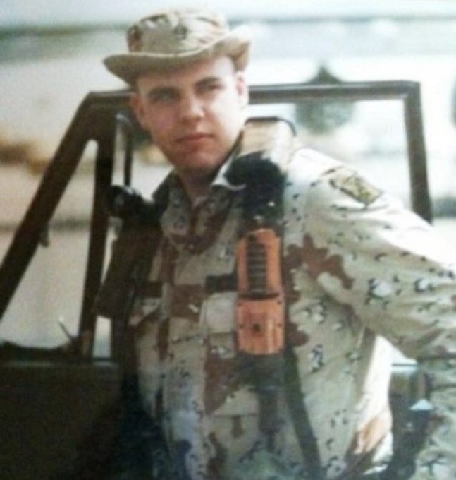 A young soldier in desert camouflage uniform and cap stands in front of a vehicle, wearing a tactical vest with a radio.