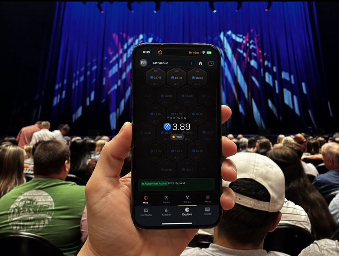 A hand holds a smartphone displaying the SatRush.io app with a wallet balance of 3.89 USD and a hexagonal grid of bitcoin values in the background, while in the blurred background a person wears a green "Epoch" shirt at what appears to be a Bitcoin conference. The image illustrates the post's message about mobile sats accumulation with SatRush.io during Epoch event.