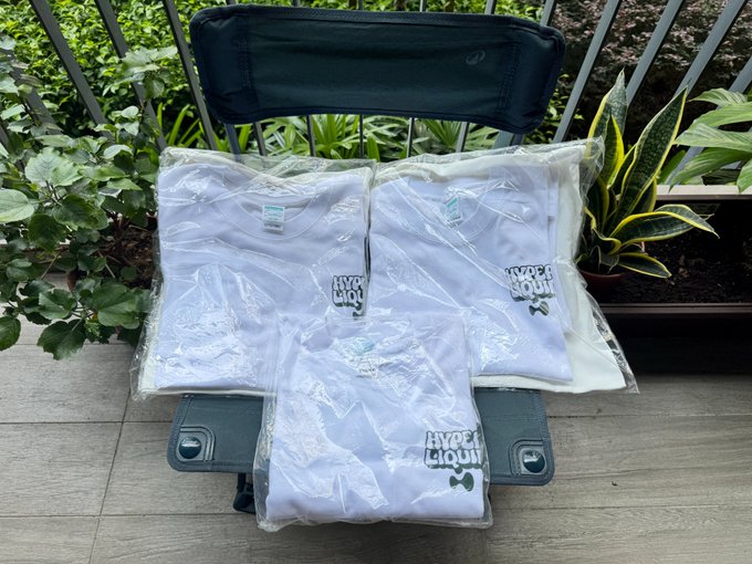 Several folded white t-shirts with 'HYPER LIQUID' branding in plastic packaging arranged on a green folding chair on a balcony.