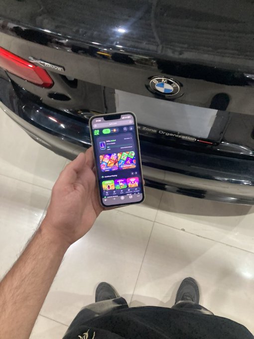 A person holds up their smartphone displaying a colorful mobile game lobby screen in front of the rear of a black BMW, likely showcasing their new car paired with gaming on the go in a "Gn bros 🖤" goodnight post.