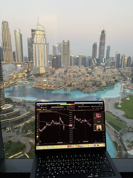 An open laptop displaying stock charts sits on a windowsill overlooking a city skyline with skyscrapers and a waterfront.
