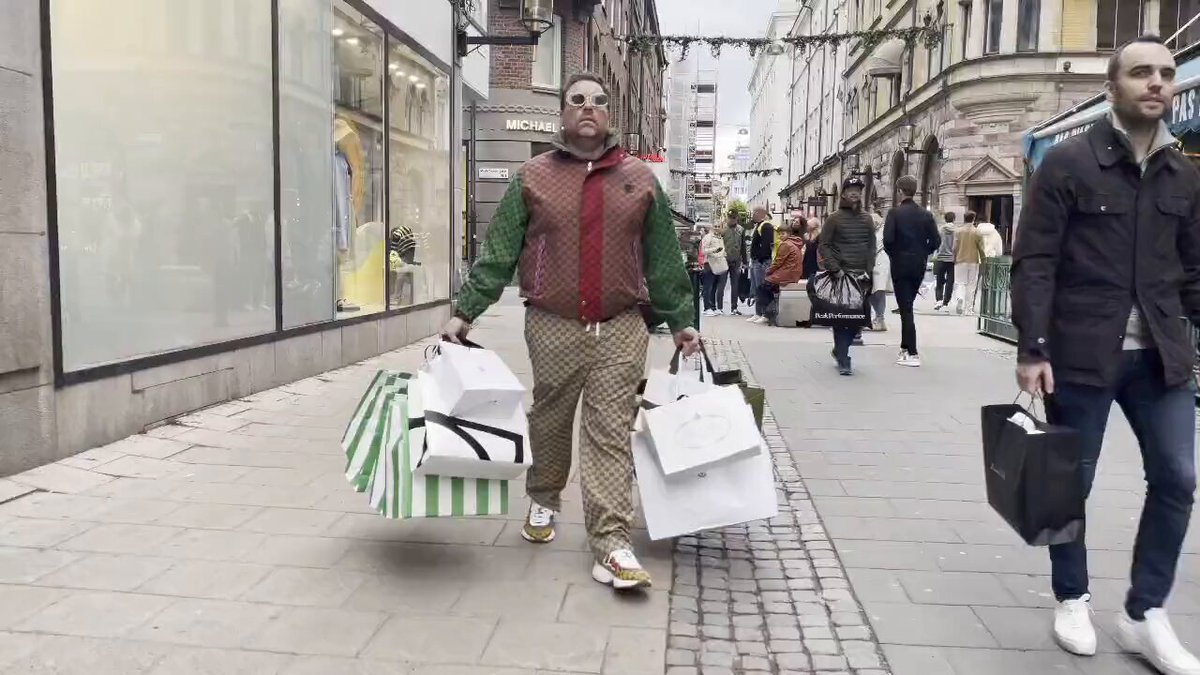 The image shows a man walking down a city street carrying two shopping bags while another person walks behind him. The scene takes place outside of a store, with people going about their day. There are also several other individuals visible in the background, some of whom may be carrying handbags or backpacks as well.