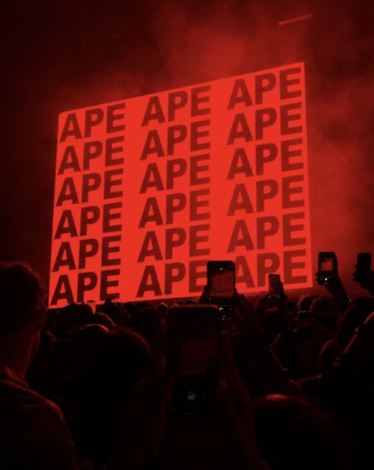 A concert scene bathed in red lighting features a large screen displaying the word 'APE' repeated in a grid pattern, with a silhouetted crowd holding up phones in the foreground.