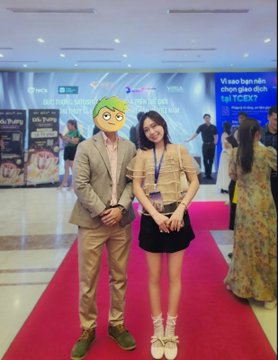 Two people posing on a red carpet at an indoor event, one wearing a green frog mask and tan suit, the other in a plaid jacket and black skirt, with a banner reading 'tại FCEK7' visible behind them.