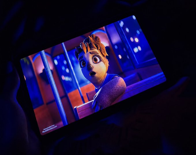 A phone screen in a dark room displays an animated scene of a worried-looking character from "The Wild Robot" movie, reflecting the cozy, indoor movie-watching vibe the post describes. The watermark reads "Official Red Carpet Team @DutchMoviesCom," fitting the theme of enjoying films at home instead of going outside.