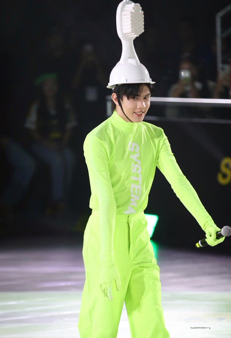 A young man wearing a bright neon green outfit with "SYSTEM" printed on it and a white helmet shaped like a toothbrush, holds a microphone on what appears to be a stage or court.