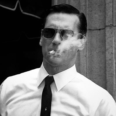 A black and white photo of a man wearing sunglasses, a white dress shirt, and a dark tie, exhaling a cloud of smoke.