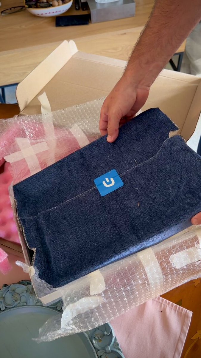 The image shows a person holding an open box containing a blue item with a white sticker, possibly a piece of artwork or collectible item. The person's hand is reaching into the box, indicating that they are about to take out the item from its packaging. The background features a wooden desk and a pink object on it.