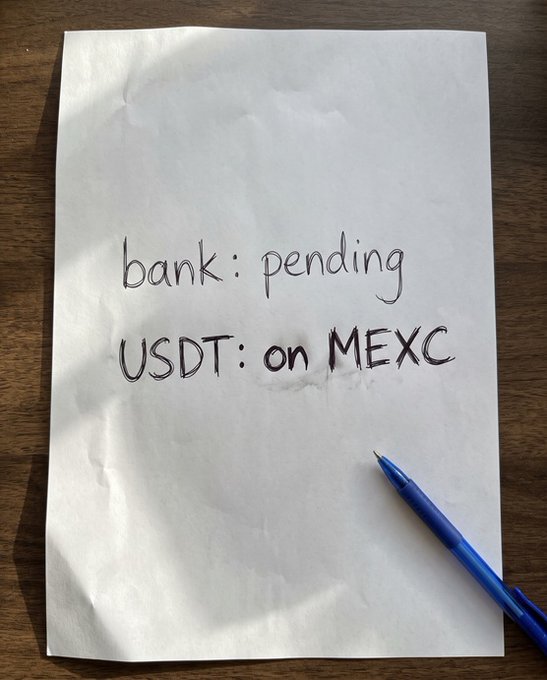 A handwritten note on white paper reads "bank: pending" and "USDT: on MEXC," with a blue pen resting beside it on a wooden surface, visually summarizing the post's point about waiting on a brokerage transfer while USDT had already arrived on MEXC for faster trading.
