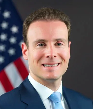 A formal portrait shows a smiling man with short brown hair, wearing a dark navy suit, white shirt, and light blue tie, posed in front of an American flag. The image accompanies a quote from CFTC Chair Rostin Behnam (Selig) advocating for Bitcoin self-custody rights, aligning with his public support for the crypto industry's foundational principles.