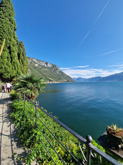 A scenic view shows a clear blue lake beside a paved walkway lined with a metal railing, lush greenery, and a palm plant, with mountains visible in the distance.