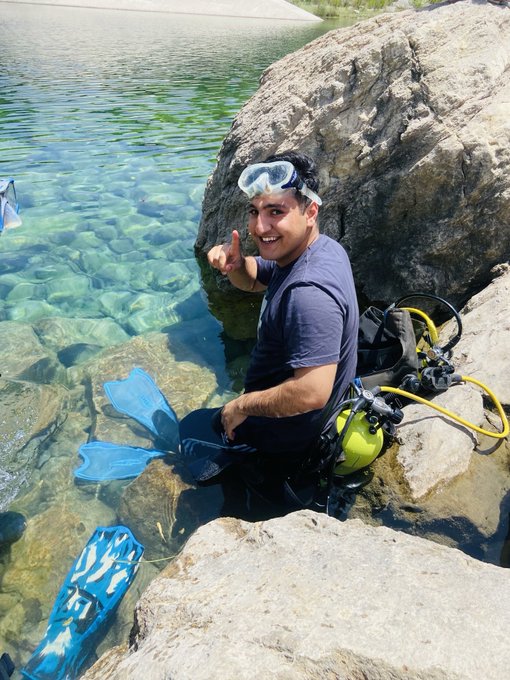 A smiling scuba diver with goggles and fins sits on a rocky shore, giving a thumbs-up next to crystal-clear water—ready to wish everyone a "GOOD MORNING" before diving in. The image captures the cheerful, adventurous spirit of the "GM" greeting, pairing the diver's enthusiastic pose with a perfect start to the day.