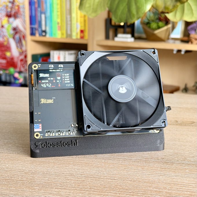 A custom Bitaxe Bitcoin solo miner (displaying "Bitaxe" branding, a hashrate of 469.2 GH/s, and 1.11% on its small screen) sits on a 3D-printed stand labeled "Solosatoshi," paired with a large computer cooling fan, illustrating the affordable, low-power home mining setup referenced in the post about running a small lottery miner for a chance at the 3.125 BTC block reward.
