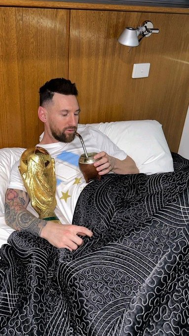 Lionel Messi rests in bed holding the FIFA World Cup trophy and drinking mate, embodying the message of rest and reset before returning stronger.