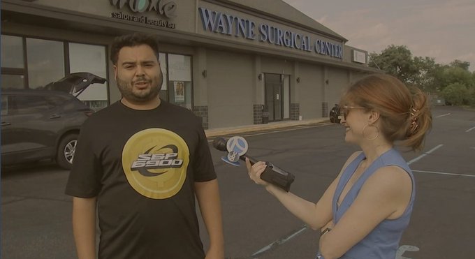 A man wearing a black 'Jet 2500' t-shirt being interviewed outside Wayne Surgical Center by a woman in a blue top holding a Channel 4 microphone, with a damaged gray car visible behind them.