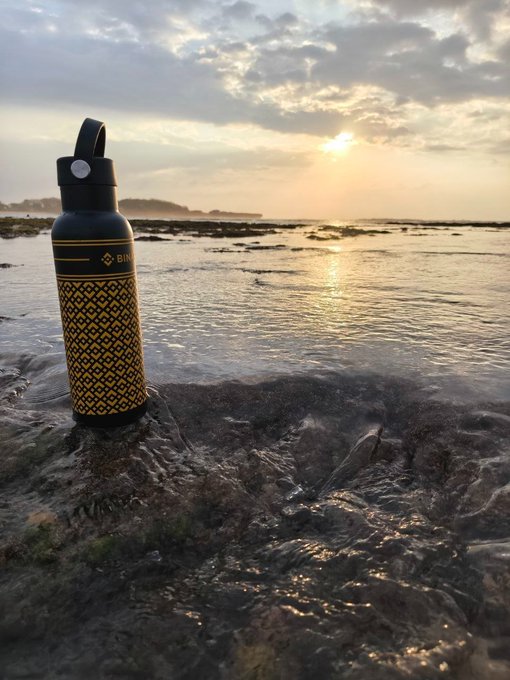 A patterned reusable water bottle stands on wet sand as the sun sets over the ocean, its golden horizon reflecting on the tide pools below. The bottle, branded "BINANCE," pairs perfectly with the post's "vitamin sea & Bⁱⁿᵃⁿᶜᵉ" pun—blending coastal serenity with a crypto lifestyle vibe.