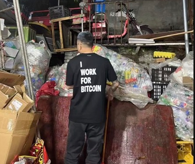 The image features a man wearing a black shirt with white text that reads "I work for Bitcoin", standing next to a pile of garbage bags, which are filled with various items including plastic bottles and cardboard boxes. The surrounding area appears to be a junkyard or storage space where discarded items have been piled up.