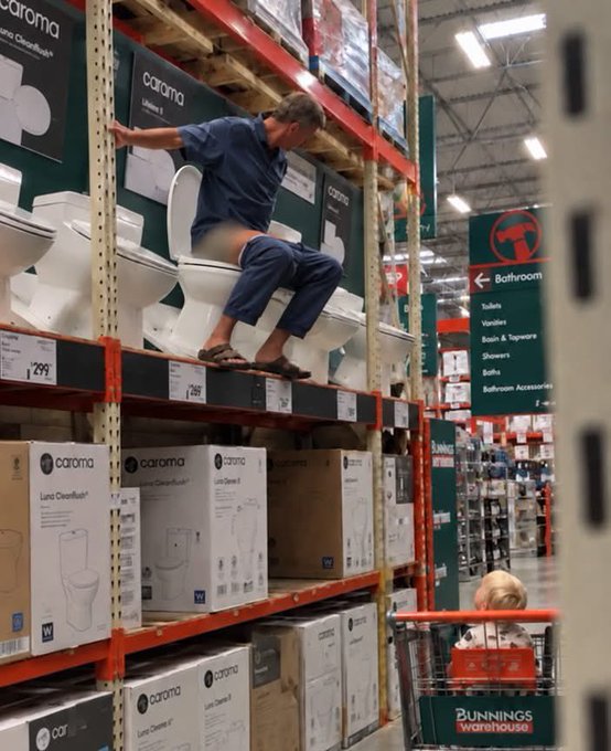The same Bunnings Warehouse scene showing a man sitting pants-down on a display toilet while a child watches from a shopping cart.