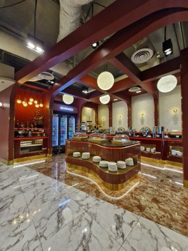 An interior view of an elegant café or restaurant featuring marble floors, red-painted wooden beams, hanging pendant lights, and a curved wooden bar with stools.