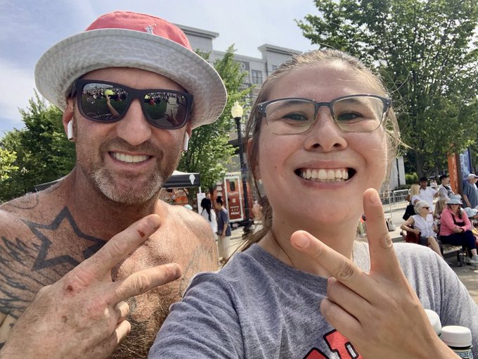 A selfie of a smiling shirtless man wearing a red cap, sunglasses, and earbuds, and a smiling woman with glasses, both making peace signs outdoors at what appears to be an event.