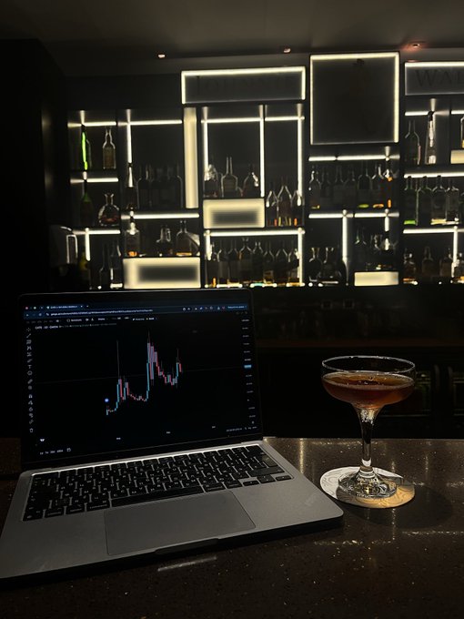 A laptop displaying a candlestick chart sits on a bar counter next to a coupe cocktail, with a dimly lit, backlit liquor display in the background—capturing the "hotel bar trading" moment described in the post where the poster serendipitously sat beside a crypto millionaire.