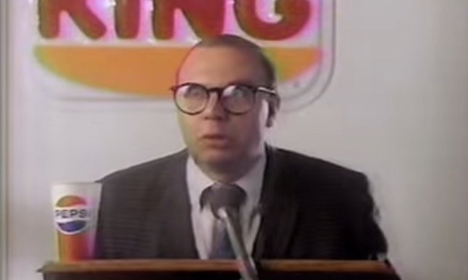 A vintage television clip showing a man in glasses and a suit at a podium with a microphone, with a Burger King logo in the background and a Pepsi cup in the foreground.