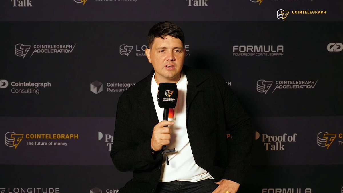 The image shows a man wearing a black jacket, seated against a white backdrop with various logos and text on it. In front of him, there's an extended microphone used for conducting interviews or live podcast sessions. The context of this scene suggests that the man is participating in a discussion, possibly at an event related to technology and AI.
