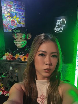 A young woman takes a selfie in a room lit with green ambient light, with a colorful painting and a neon sign visible in the background.
