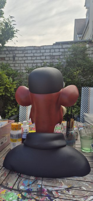 A painted sculpture of a red-headed figure in a black hat and outfit, displayed outdoors on a paint-splattered table.