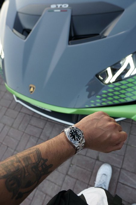 A person with a tattooed forearm wearing a stainless steel watch (appearing to be a Rolex Explorer) poses with a clenched fist in front of a matte blue Lamborghini Huracán STO with green accents. The "STO" badge is visible on the car's hood, aligning with the "GN chads" post by showcasing an ultra-rare, high-performance supercar fit for an elite lifestyle.