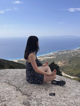A woman in a floral dress sits cross-legged on a stone overlook, gazing out at a vast landscape of sea and coastline beneath a clear blue sky.