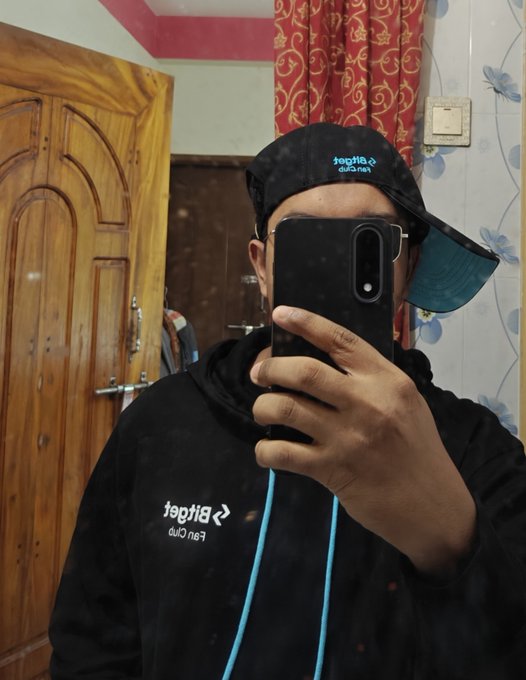 A person wearing a black cap and hoodie takes a mirror selfie with a black smartphone in a room with wooden doors and red curtains.