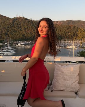 A woman with curly hair wearing a red dress poses on an outdoor seating area with a marina, boats, and a hillside in the background.