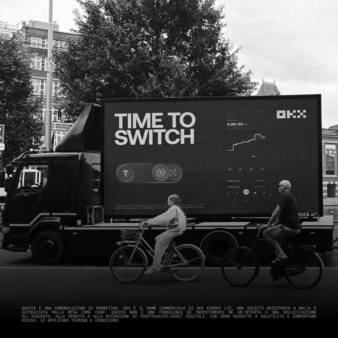 A black truck with a large digital billboard displaying 'TIME TO SWITCH' and the OKX logo is parked on a city street as a man and woman ride bicycles past it, with Italian regulatory text at the bottom.