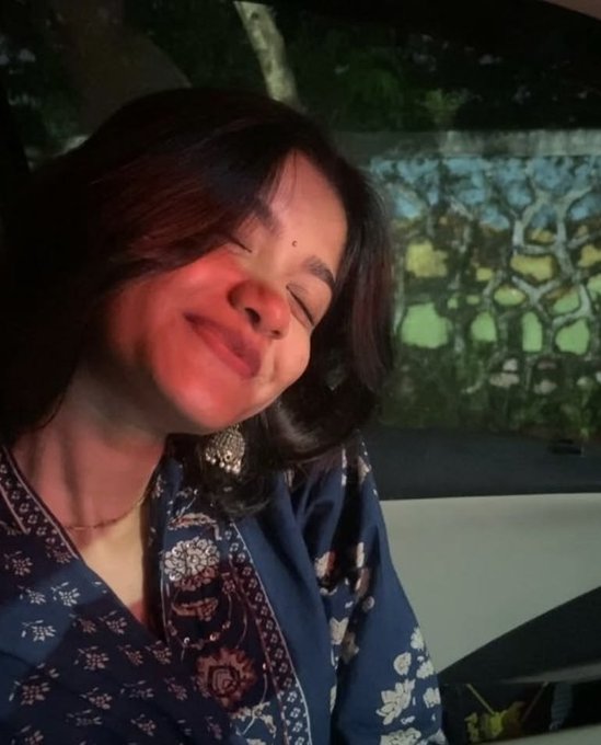 A woman with shoulder-length dark hair smiles warmly with closed eyes while wearing a dark blue patterned top and a silver earring.