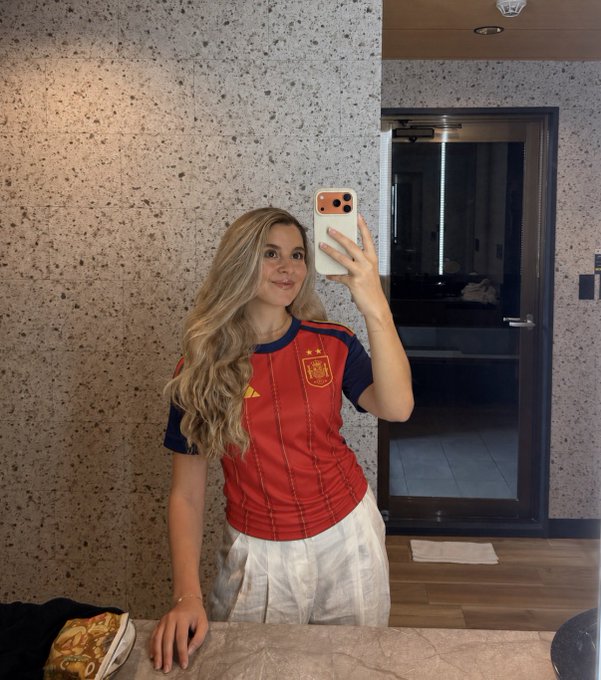 A blonde woman takes a mirror selfie wearing a red Spain national football team jersey with the team's crest and Adidas logo visible, paired with white pants. The image matches the celebratory post about Spain's victory, with her posing confidently to show off the team colors.