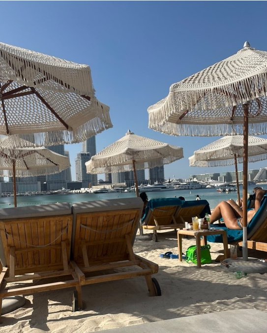A scenic beach view with white fringed umbrellas, wooden lounge chairs, people relaxing, and a city skyline with yachts in the background.