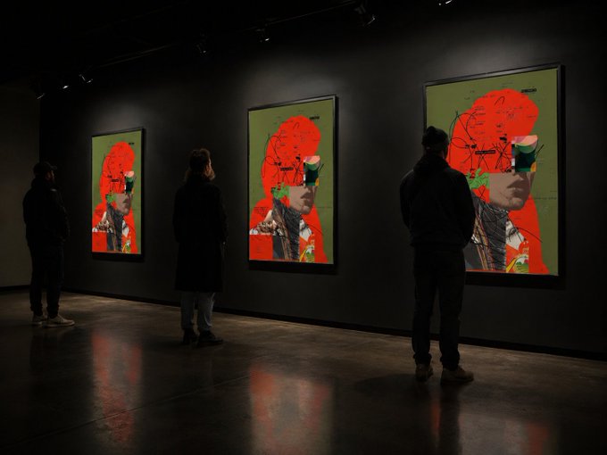 Three people viewing large, identical, vibrant green and red portraits displayed on the dark walls of a modern art gallery.