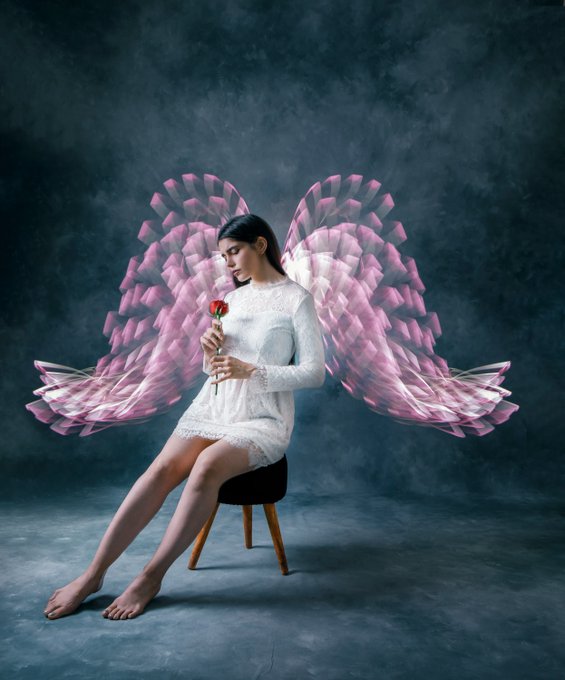 A young woman in a white lace dress sits barefoot on a stool holding a single red rose, with large, elaborate pink angel wings emerging from her back against a dark, smoky background.
