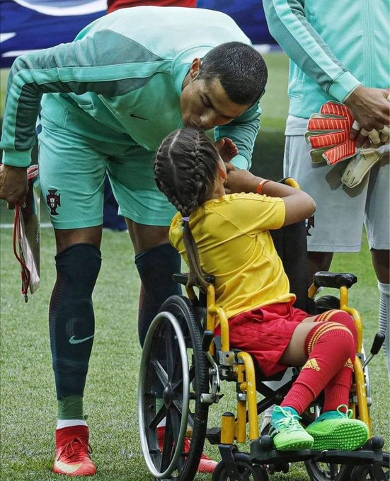 A soccer player in a light green Portugal national team uniform (with Nike and Portuguese crest visible) bends down to embrace a young child in a yellow shirt seated in a yellow wheelchair on a field.