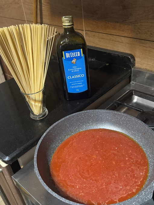 A bottle of DeCecco Classico extra virgin olive oil sits next to uncooked spaghetti in a glass and a pan of red pasta sauce on a stovetop.