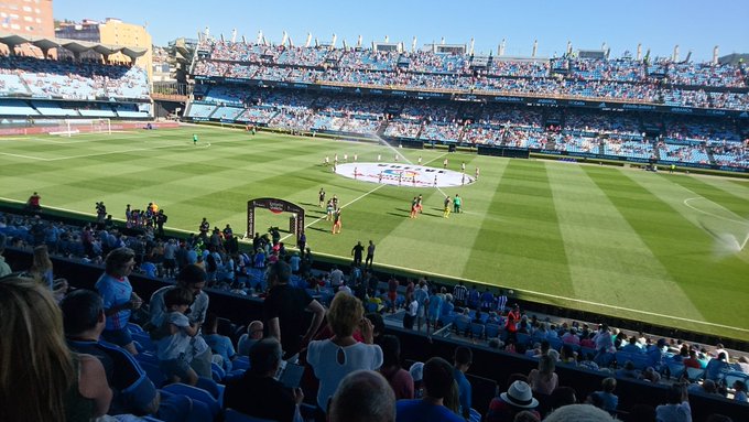 A soccer/football stadium with a large crowd watching a match on a sunny day, taken from the stands.