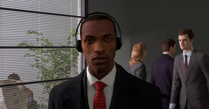 A screenshot from a video game (likely Grand Theft Auto) showing a man in a black suit, red tie, and headphones standing in an office with other people in suits in the background.