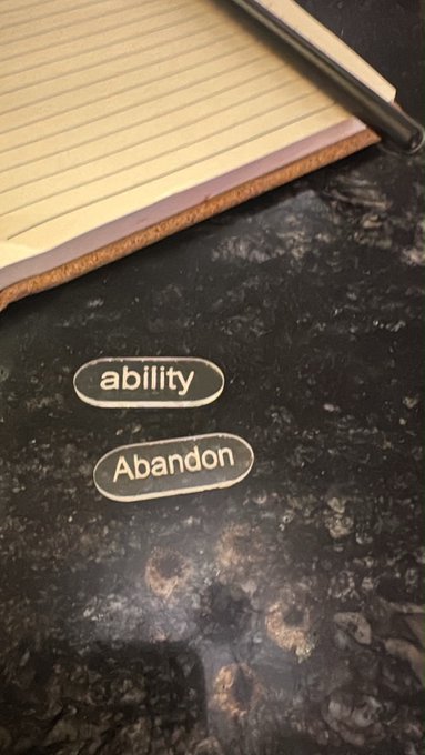 A photograph showing a notepad and pen on a dark granite countertop, with two oval word stickers labeled 'ability' and 'Abandon' placed on the surface.