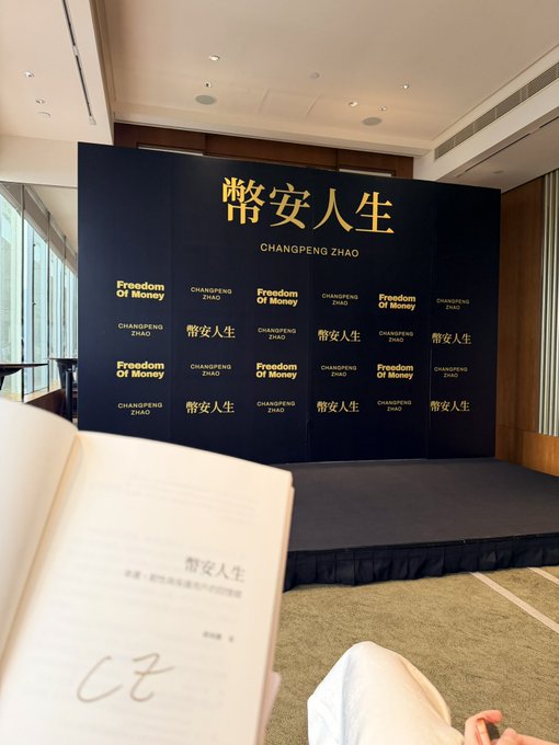 A reader holds a copy of Changpeng Zhao's book "幣安人生" (Freedom of Money) in the foreground, while a backdrop displays the book's title and CZ's name, fitting the BNB KOL AMA context with CZ.