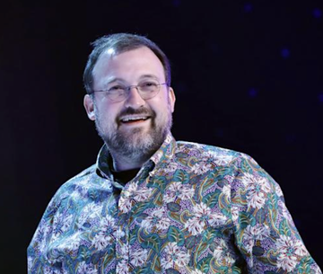 The image features a man wearing glasses, smiling at the camera with his hands on his hips. He is dressed in a shirt adorned with white flowers. The background of the image is dark blue, which contrasts with the man's attire. In the bottom right corner of the photo, there is text that reads "NEW: Charles Hoskinson is launching a new Discord hub for Cardano discussions". This suggests that Charles Hoskinson has created a platform specifically designed for cardano enthusiasts to engage in discussions and share information about the Cardano cryptocurrency.
